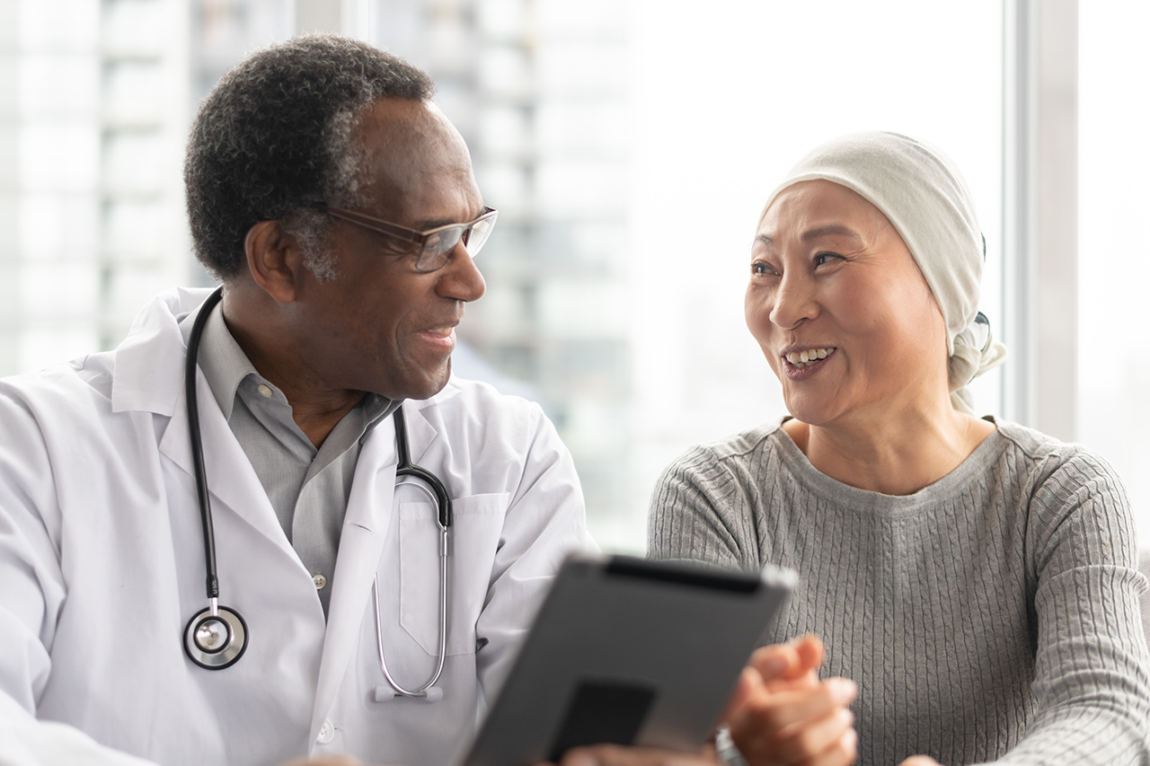 A Korean woman with cancer is meeting with her African American male doctor. The doctor is showing her test results on an electronic wireless tablet. The doctor smiles as he gives good news regarding the patients treatment