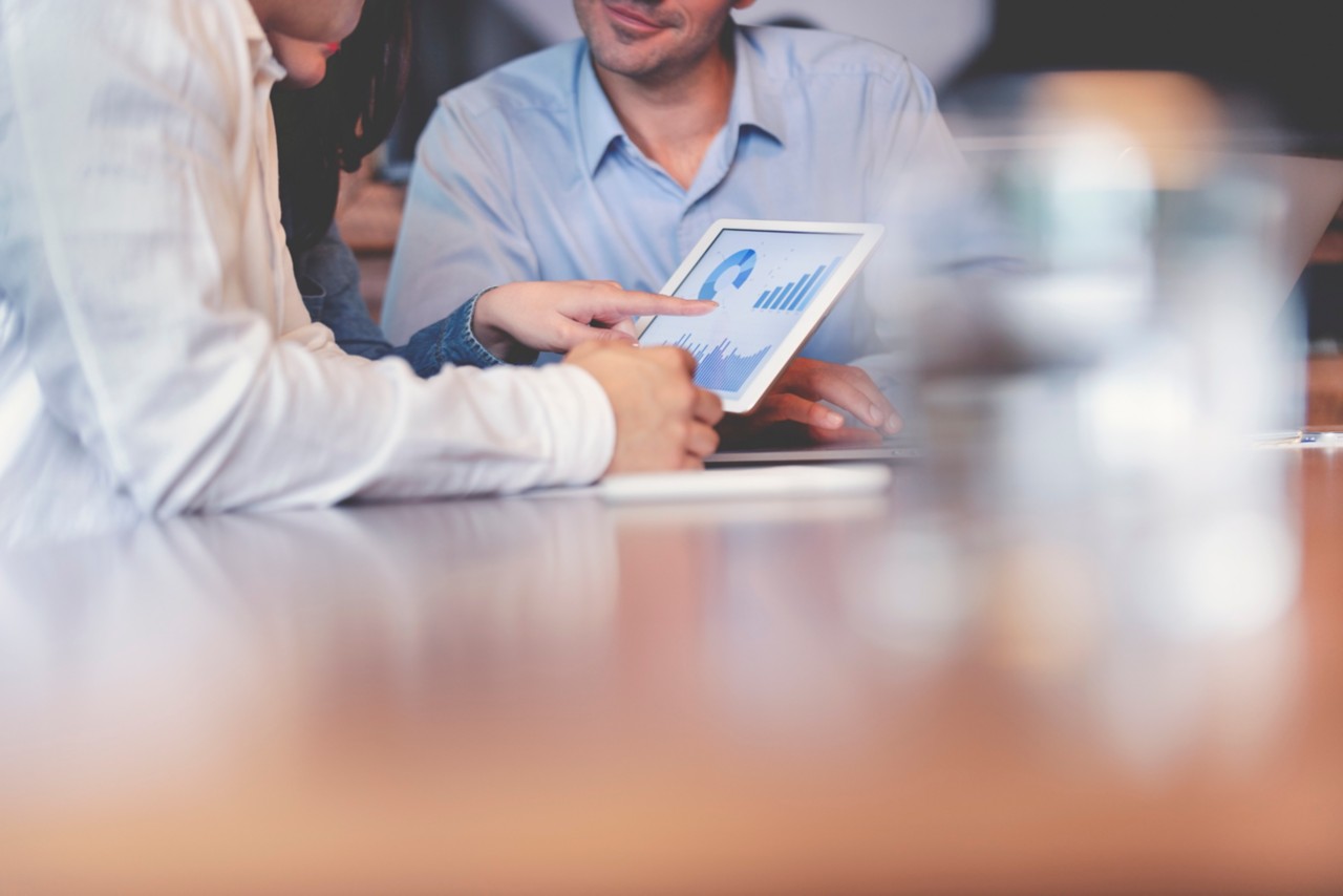 Business people working on financial data on a digital tablet. One looks like the manager working with employees, or a financial advisor. It may also be a training session. All are dressed in business casual clothing.