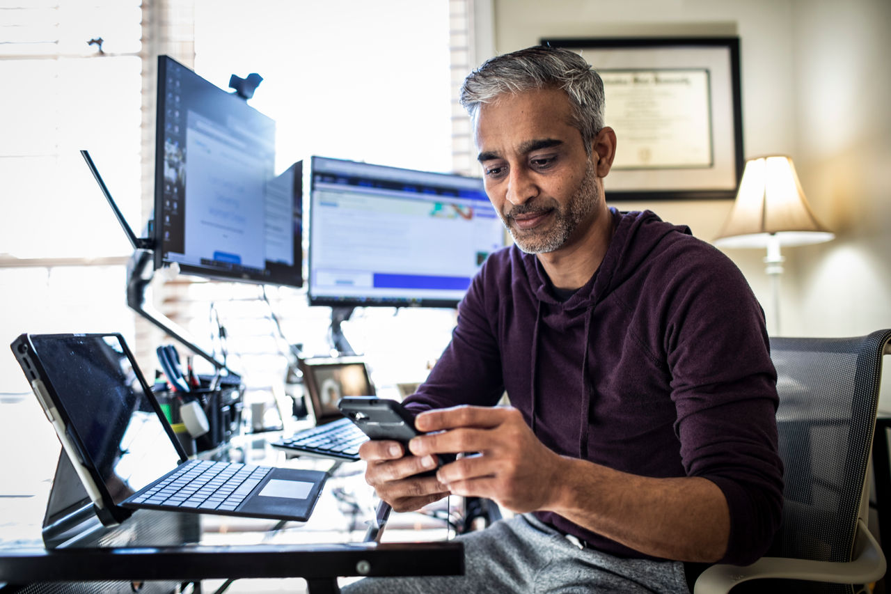 Man working in home office at desk with computers looking at mobile phone