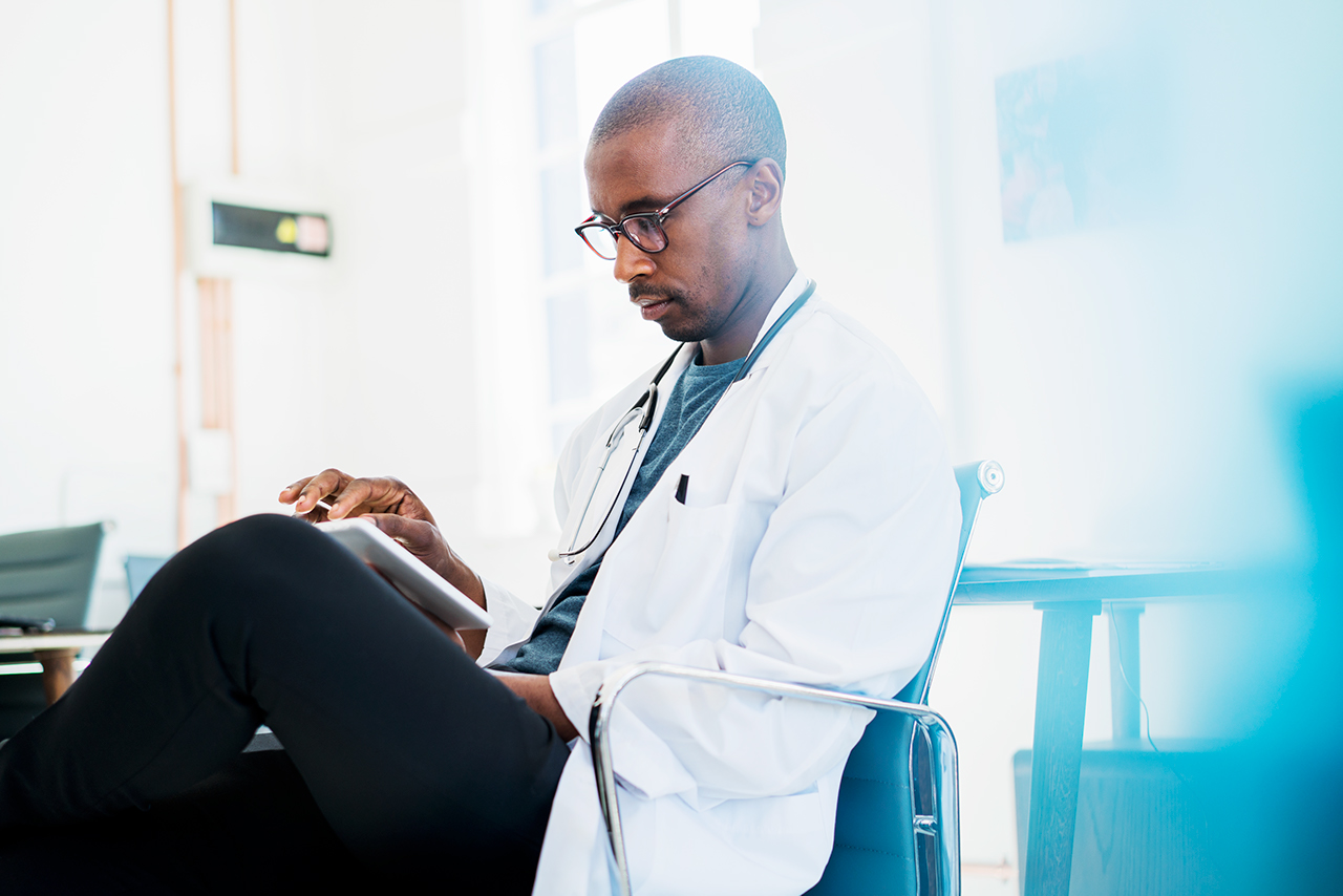 A photo of doctor using digital tablet. Concentrated male professional sitting on chair. He is brightly lit hospital.