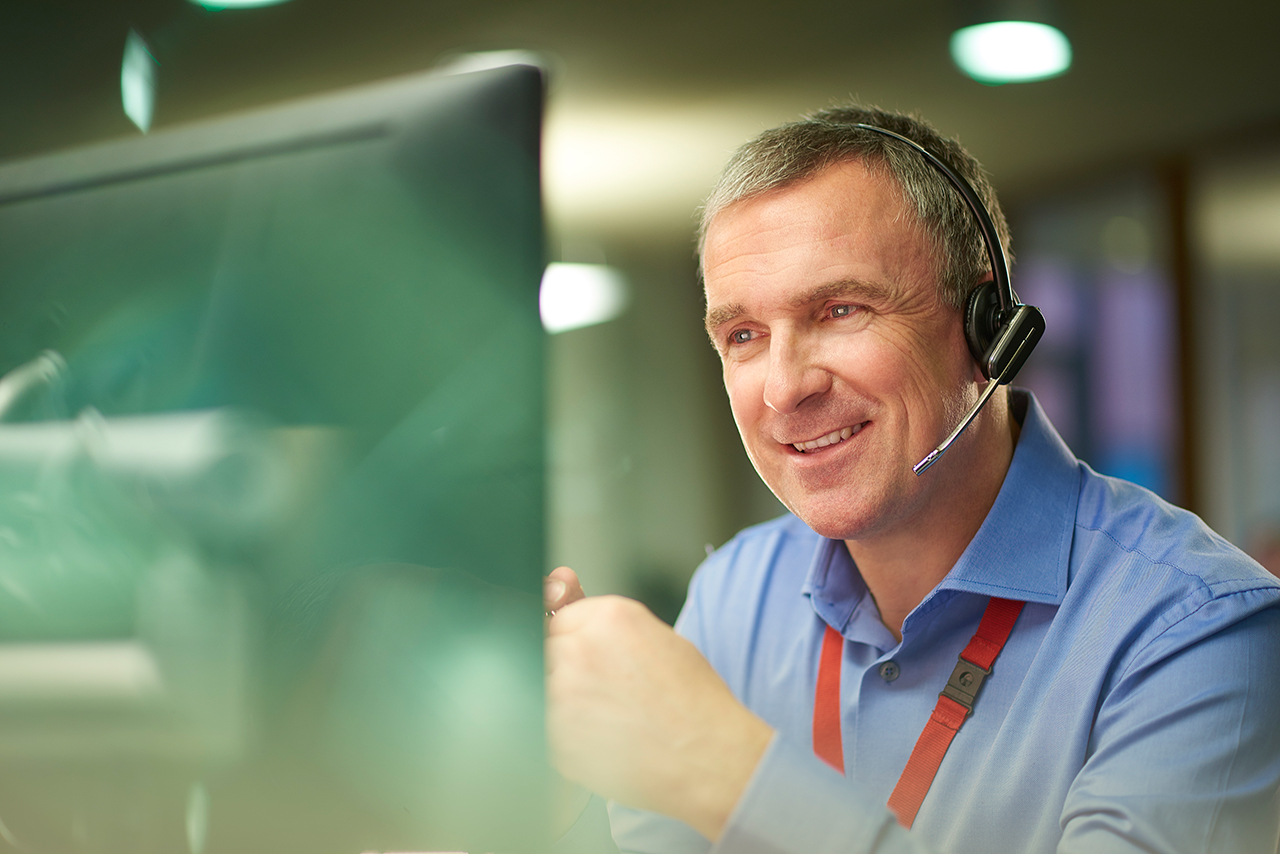 a call centre phone operative in his mid 40s chats on the phone at his desk . He is explaining something to the person on the phone in a friendly manner . behind him a defocussed office interior can be seen . This could be a call centre or an office worker chatting to a customer .