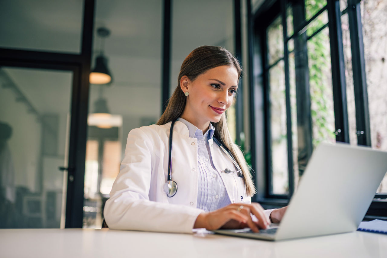 Positive doctor working on laptop in medical office, portrait.